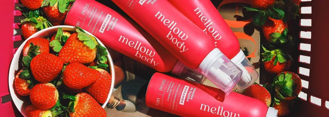 Imagem vibrante com frascos do mousse de banho "mellow body" sabor morango, cercados por morangos frescos em uma tigela e ao redor, transmitindo frescor, doçura e cuidado pessoal.